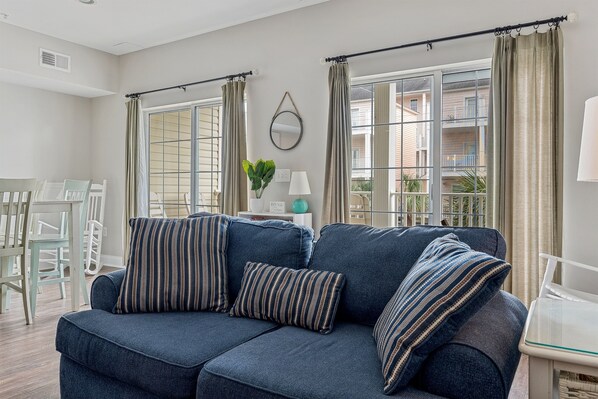 Un salon avec un canapé bleu foncé, deux coussins à rayures, un miroir rond accroché au mur et une vue sur un balcon avec des immeubles en arrière-plan.