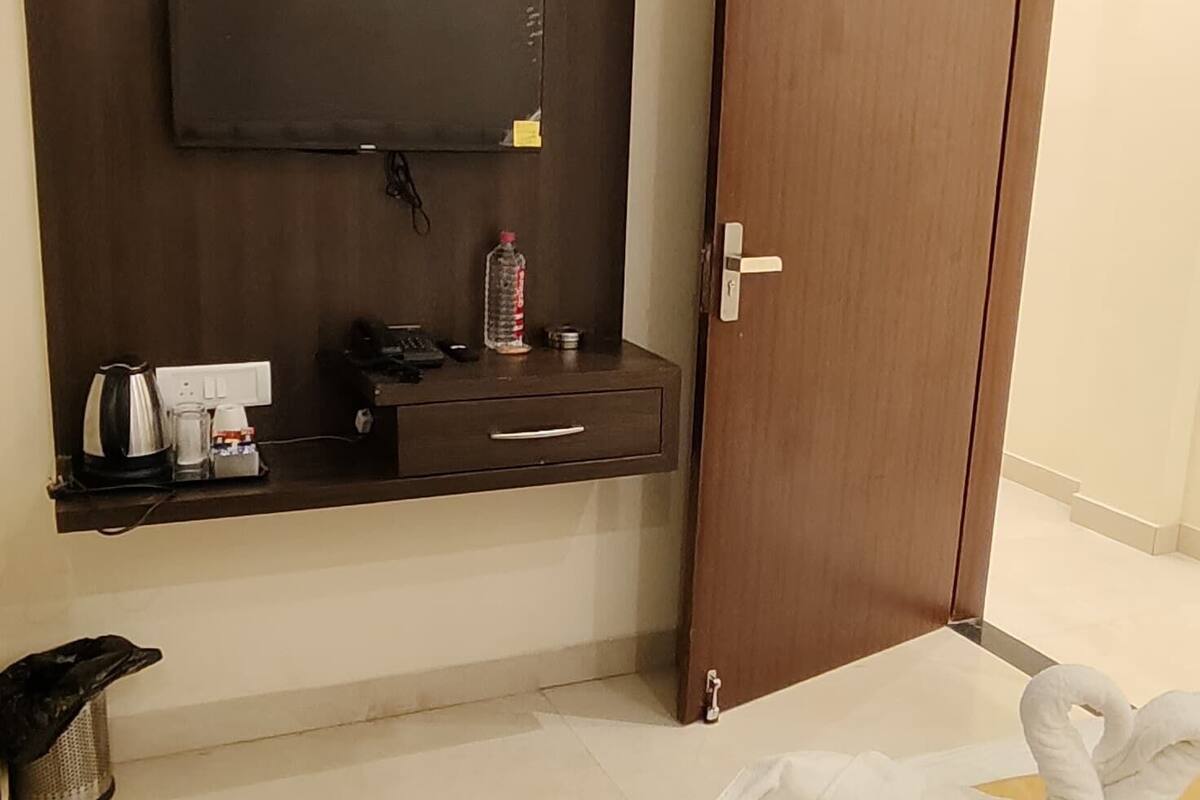 A hotel room with a bed, a TV mounted on the wall, a wooden door, and a small table with a kettle and a bottle.