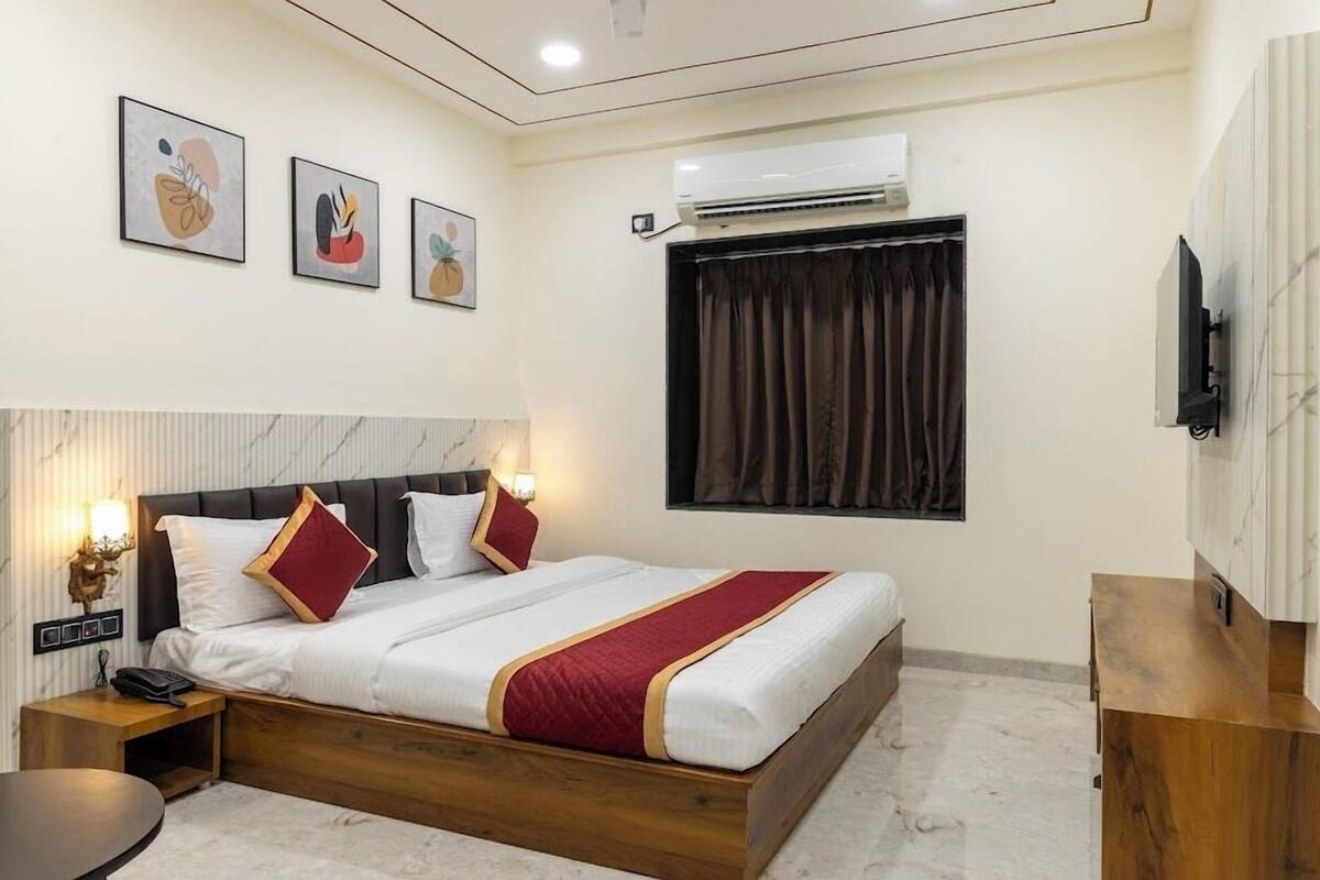 A hotel room with a bed, bedside table, lamp, artwork, air conditioning unit, and a TV mounted on the wall.