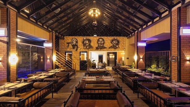 A restaurant with a rustic interior, featuring exposed brick walls, wooden beams, and a central staircase. Tables are set with plates and glasses, and there are plants and a chandelier visible. - Hotel Money Plant (Chhatrapati Sambhajinagar)