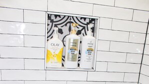 Een badkamer met witte metrotegels, een zwarte douchebank en een plank met drie flessen Pantene Moisture Repair Shine en Olay Moisturizing Repair Shine.