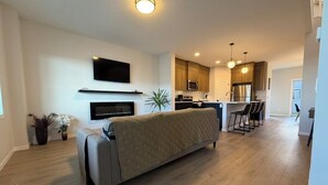 A modern living room with a sofa, a flat-screen TV mounted on the wall, a kitchen area with wooden cabinets and a microwave, and a dining area with bar stools.