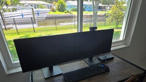 A computer desk with a large monitor, keyboard, and mouse, positioned near a window with a view of a fenced outdoor area and buildings. - Gulfside Getaway - Newly renovated waterfront 3-bedroom w/dock in Port Richey (Port Richey)