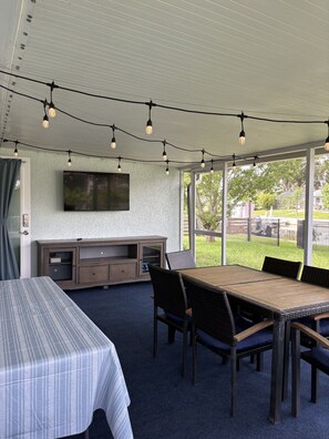 An outdoor covered patio with a dining table and chairs, a TV mounted on the wall, and string lights overhead.