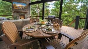Une terrasse en bois avec une table ronde dressée pour le repas, entourée de chaises, et une cheminée en pierre.