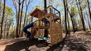 A wooden play structure in a forest with a slide and climbing wall.