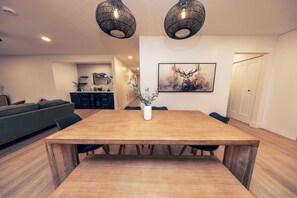 Un coin repas avec une table en bois, deux luminaires suspendus, un vase avec des plantes et une œuvre d’art encadrée au mur.
