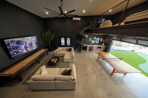 A modern living room with a large flat-screen TV, a sofa, a coffee table, and a dining area with a bench.