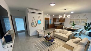 En moderne stue med sofa, sofabord og dekorative genstande. Der er et køkkenområde med skabe og et spisebord.