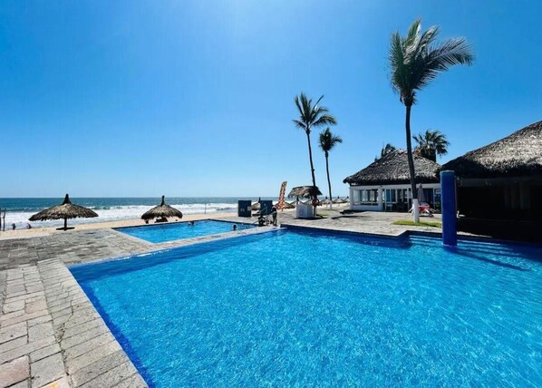 A clear blue pool with a view of the ocean, surrounded by palm trees and thatched-roof structures.