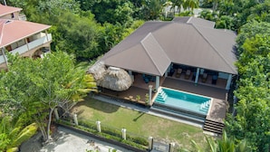 Una casa de dos pisos con piscina, rodeada de exuberante vegetación y una estructura de techo de paja.