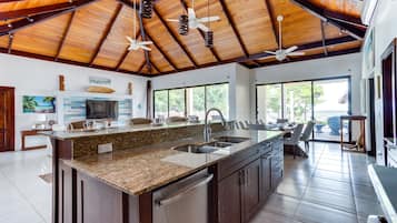 A spacious kitchen with a large island, granite countertop, and stainless steel appliances. There are ceiling fans, large windows, and a view of the outdoors.