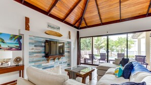 Un salón con televisión en la pared, un sofá, una mesa de centro y vistas al exterior a través de amplias ventanas.