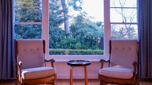 A room with two wooden chairs and a small round table, large windows with a view of greenery outside. - Sublime Art Deco Cottage: Leura, Blue Mountains (Leura)