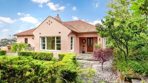 A two-story house with a tiled roof, a front porch, and a well-maintained garden with various plants and a small tree. - Sublime Art Deco Cottage: Leura, Blue Mountains (Leura)