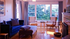 A cozy living room with a blue sofa, a wooden coffee table, and two armchairs. Large windows offer a view of greenery outside. - Sublime Art Deco Cottage: Leura, Blue Mountains (Leura)