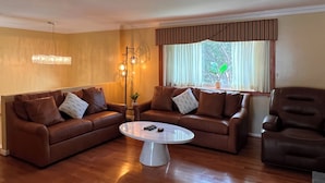 Ein Wohnzimmer mit zwei braunen Ledersofas, einem weißen Couchtisch und einem braunen Sessel. Ein Stehaufglampen und ein Blumentopf stehen auf einem Fensterbrett.