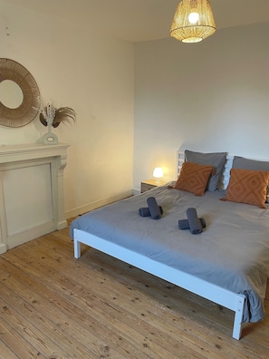 A bedroom with a white bed, wooden flooring, a bedside table with a lamp, and a mirror on the wall.