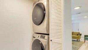 Un cuarto de lavandería con dos lavadoras apiladas, una puerta con listones horizontales y una vista parcial de una sala de estar con un sofá verde y un sillón amarillo.