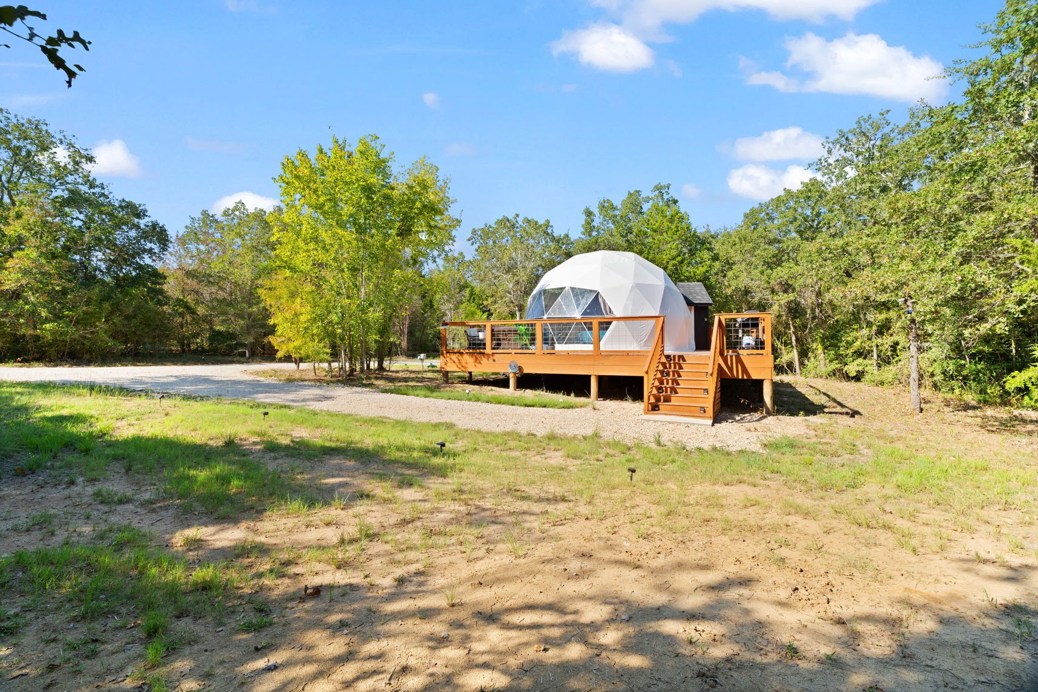 A dome-shaped cabin with a wooden deck and a ladder, surrounded by trees and a gravel path.