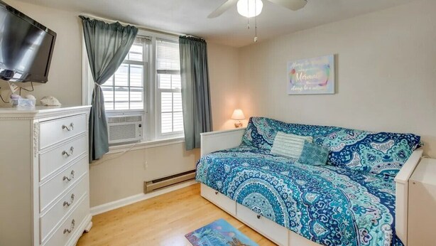 Un dormitorio con cama, cómoda, ventilador de techo, ventana con cortinas y televisor montado en la pared.