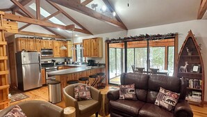 A spacious living room with a brown leather sofa, two armchairs, and a coffee table. A wooden dining table is in the center. The room has wooden beams on the ceiling and a large window with a view of trees. - Riverside Cabin Near Torch Lake – Kayaks Included! (Rapid City)