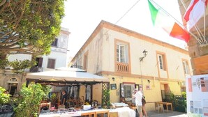 An outdoor market with tables displaying various items, a building with a balcony, and a flag in the background. - Apartments La perla - Apartment with Balcony (Carloforte)