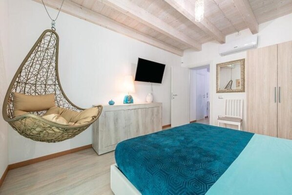 Un dormitorio con una silla colgante, una cama con colcha estampada, un televisor en la pared y un armario.