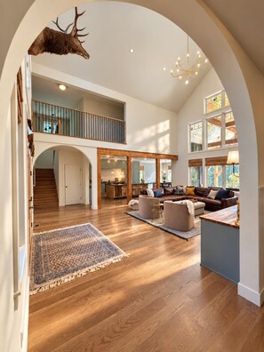 Un intérieur spacieux avec un plafond haut, un plancher en bois, un espace salon central et une cuisine visible à travers une arche.