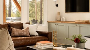 A living room with a brown leather sofa, a coffee table with books, a TV on the wall, and a window with a view of trees. - Luxury Golf Suncadia Retreat- Hot Tub + Sleeps 20 (Cle Elum)
