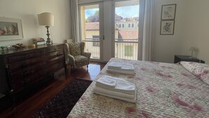 A hotel room with a bed, a dresser, a chair, and a balcony with a view of buildings. - Residence Clementina, 155sqm of charm in the historic center (Padova)