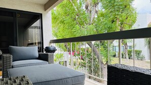 Un balcon avec un canapé et un barbecue, donnant sur un jardin agrémenté d’arbres et de plantes.
