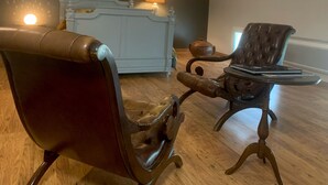 Une pièce dotée d’un parquet en bois foncé, d’un fauteuil en cuir marron, d’un pouf assorti, d’une petite table ronde sur laquelle se trouve un livre, une lampe et un meuble décoratif.