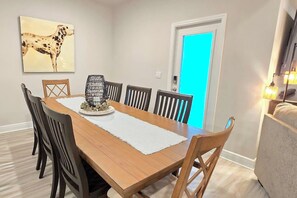 A dining room with a wooden table, six chairs, a painting of a dog, a door with a blue light, and a lamp on the right. - Dog-Friendly Beach Home · Golf Cart · Beach Access (Port St Joe)