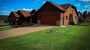 A large, two-story wooden house with a garage, a covered driveway, and a well-maintained lawn. - Serene Golf Course Home Getaway with Hot Tub (Sturgis)