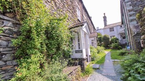 Un étroit sentier de pierres mène à une petite maison blanche portant l’inscription « Cosy Cottage 1 Tom Fold ». Un mur de pierres borde le sentier d’un côté, et de l’autre, une zone herbeuse est parsemée de plantes.