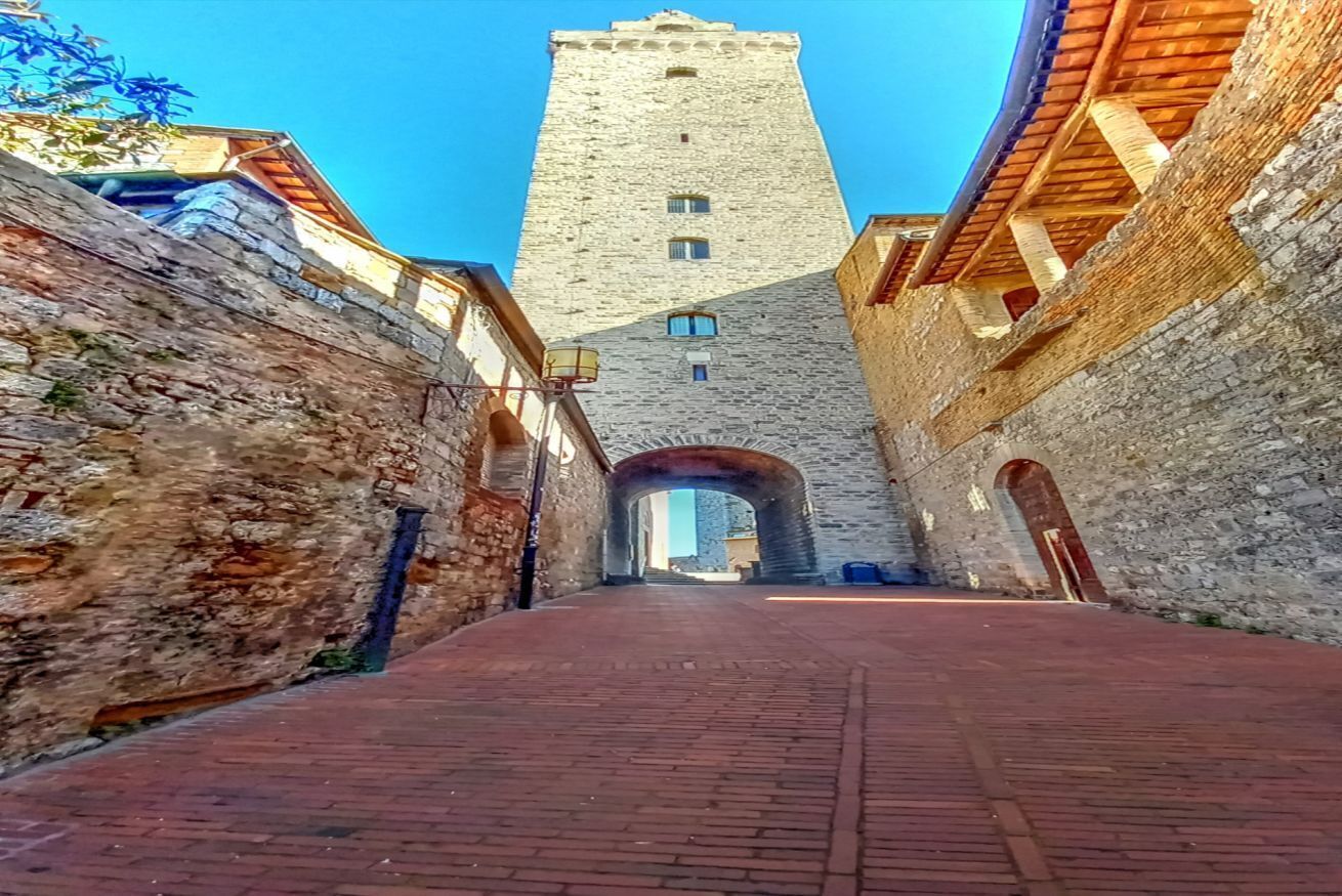A narrow alleyway with a stone tower in the distance, flanked by buildings with wooden balconies.