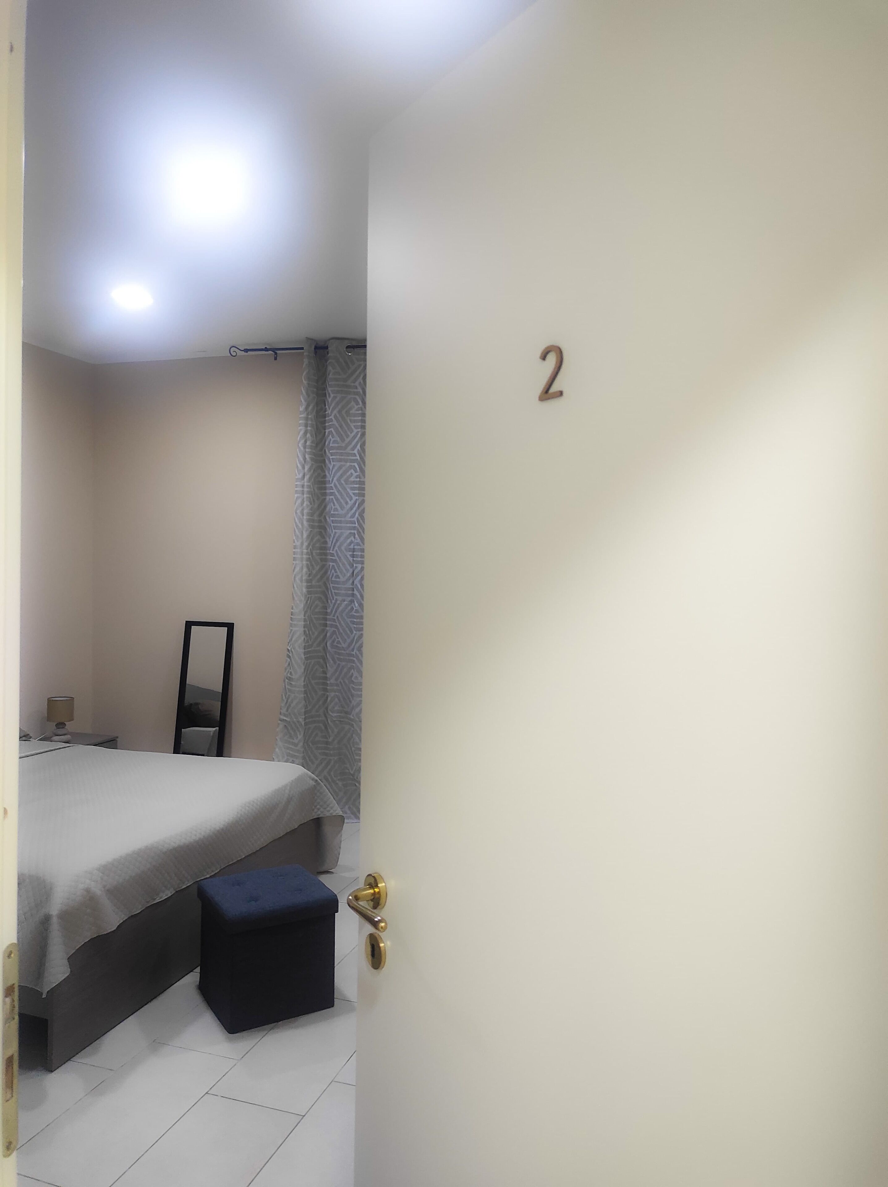 Een slaapkamer met een bed, nachtkastje, spiegel, gordijn en een deur met een nummer.
