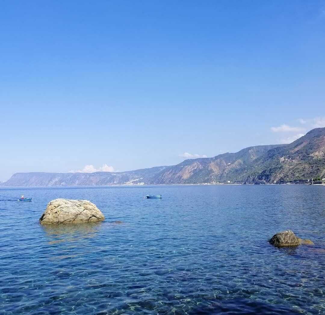 Helder uitzicht op de kust met kalm water, een grote rots op de voorgrond en een bergketen in de verte.