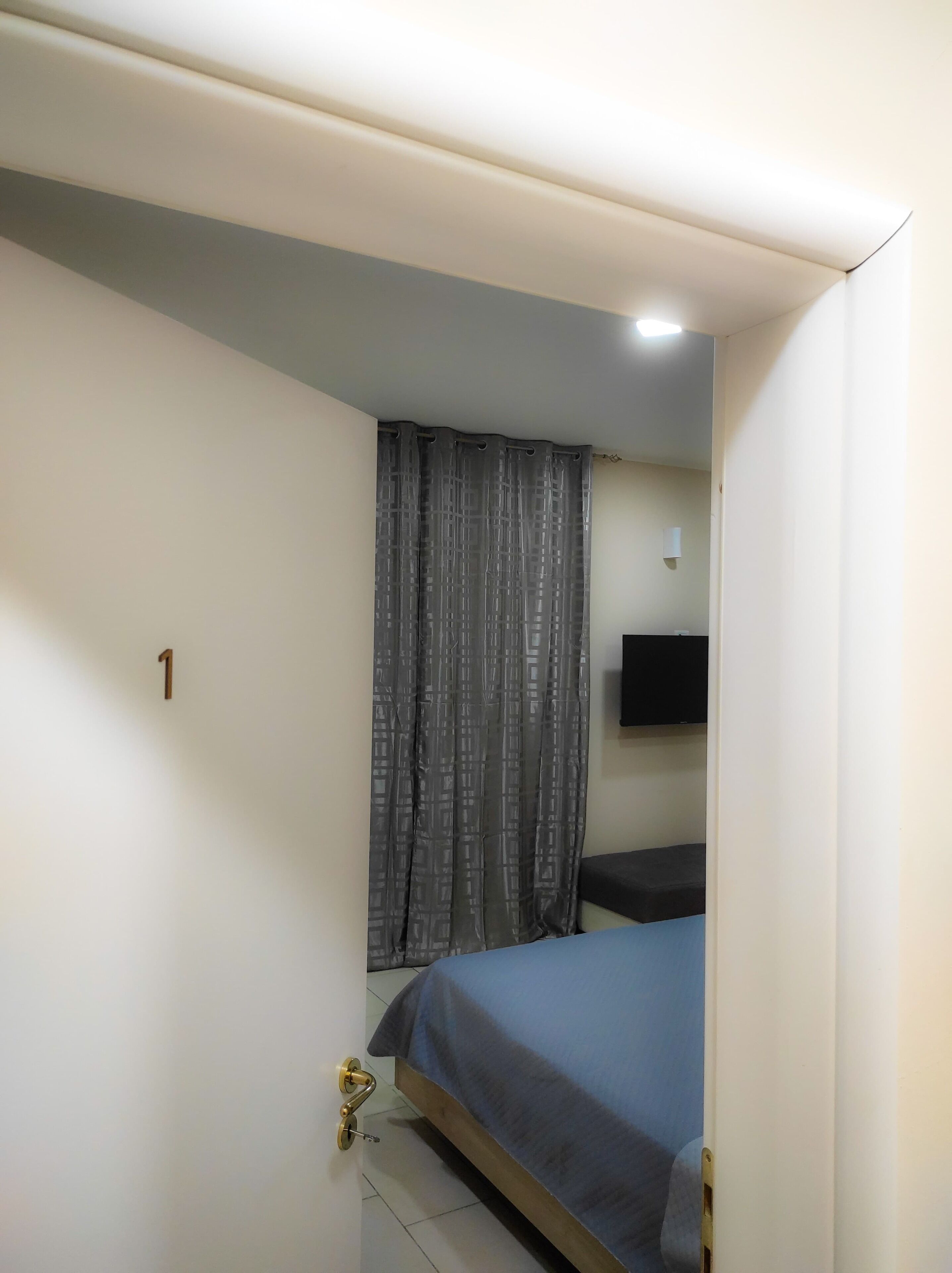 Eine kleine, einfache Unterkunft mit einem Bett, einem Vorhang, einem an der Wand befestigten Fernseher und einer Tür mit der Nummer 1.