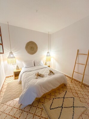 A neatly made bed with white linens, two bedside lamps, a woven wall hanging, and a patterned rug. - Eco Bay hostel  (Aourir)
