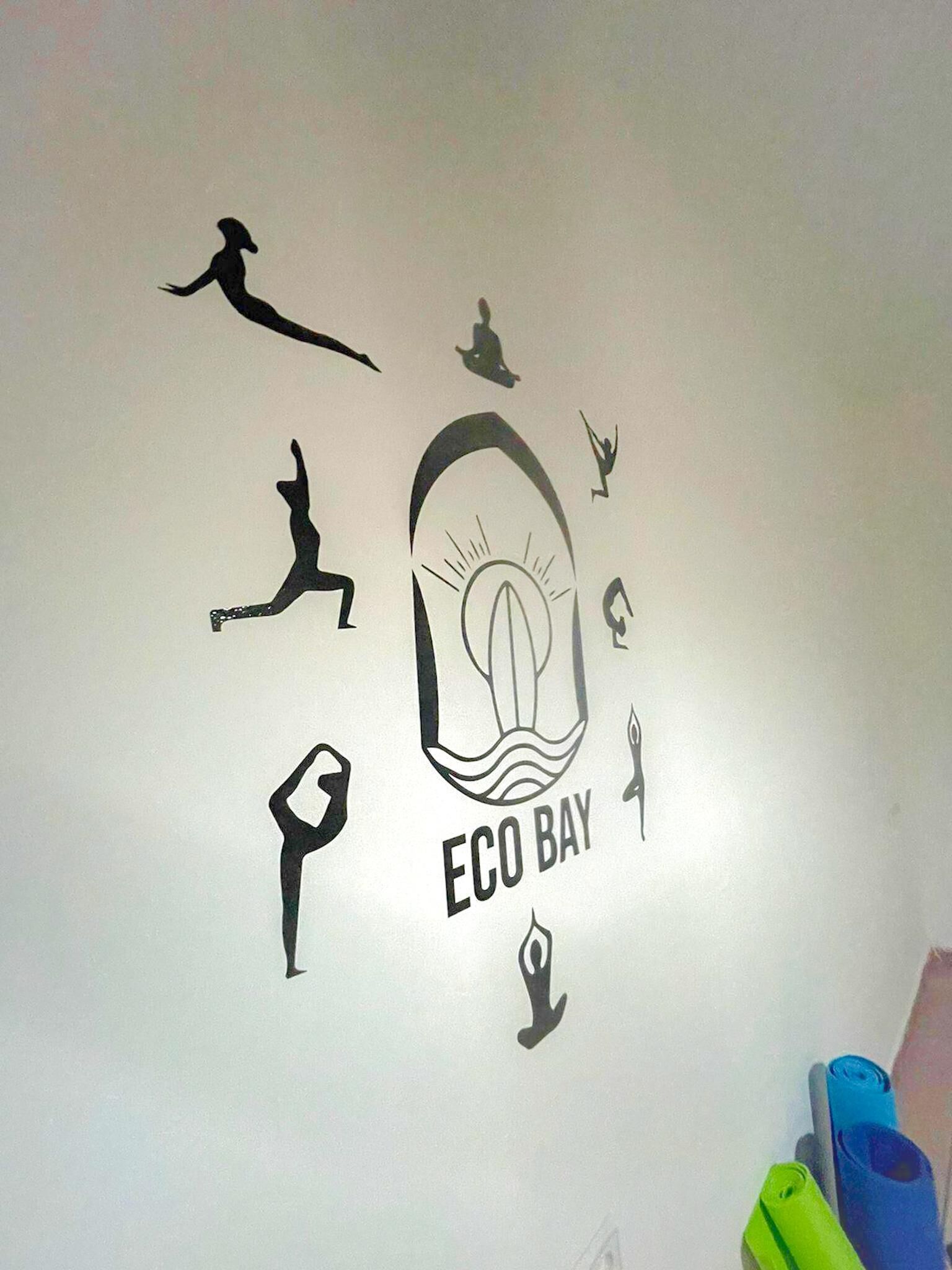 A wall with various black silhouettes, including a person in a hat, a person in a yoga pose, and a person jumping. The central image is a stylized eye with the text "ECO BAY" below it.