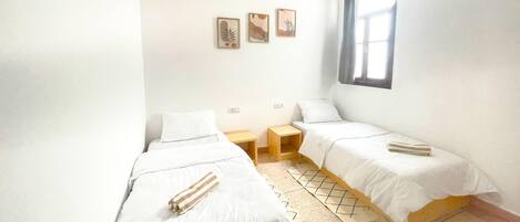 A small, bright room with two single beds, a window, and a hanging light fixture.
