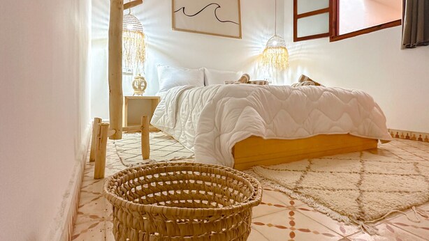 A bedroom with a bed, a woven basket, a wooden ladder, a wall-mounted picture, and a window with curtains. - Eco Bay hostel  (Aourir)