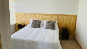 Un dormitorio con cabecera de madera, dos almohadas con motivos geométricos, una colcha blanca, una mesita de noche con control remoto y un cesto de basura pequeño.