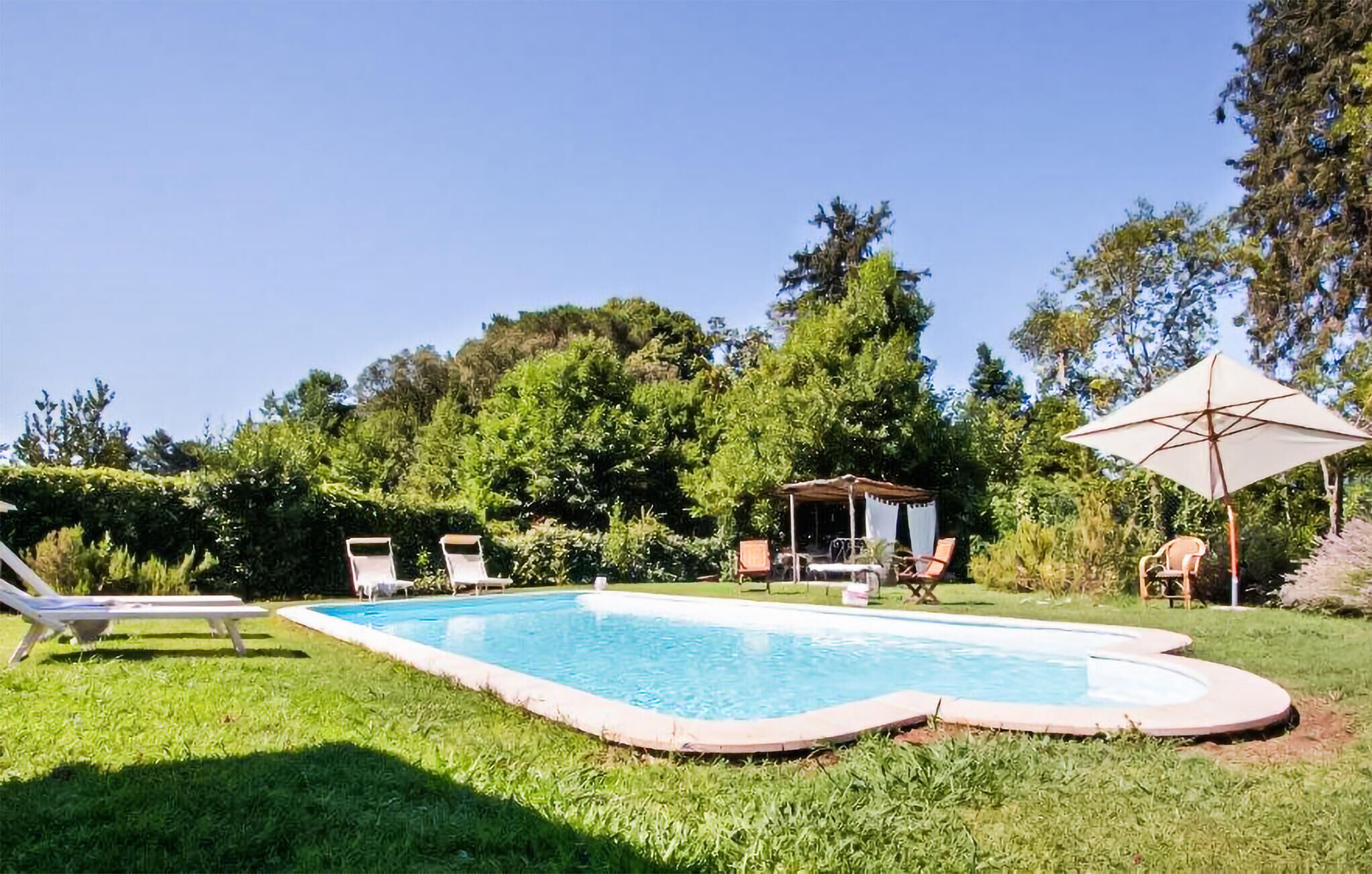 A rectangular swimming pool with a tiled edge, surrounded by a grassy lawn. There are lounge chairs and a covered seating area with a table.