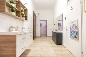 Ein Flur mit Holzboden, weißen Wänden, einer weißen Theke mit Waschbecken und einem Spiegel. Blumentöpfe stehen auf Regalen und eine Tür trägt eine Nummer.