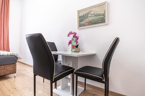 Quarto com uma mesa branca, duas cadeiras pretas com detalhes dourados, um pequeno vaso com flores e um quadro na parede.