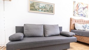 A grey sofa with two cushions, a painting of a landscape, and a bed with a patterned blanket. - Cozy Central Room, Only 15-Min Walk to Prater (Vienna)