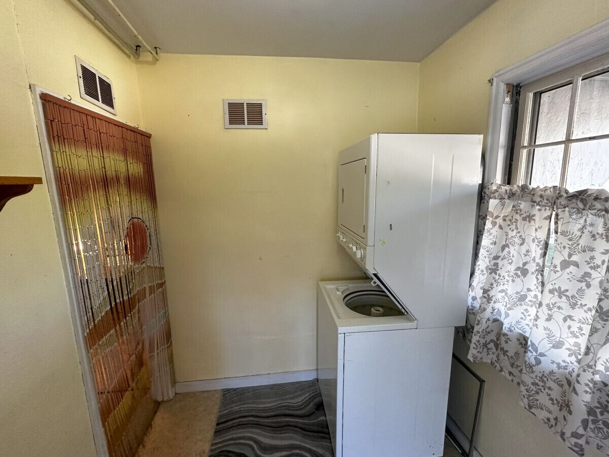 A compact kitchen with a washing machine, a small refrigerator, and a window with floral curtains.